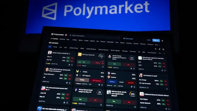 Esta fotografia mostra telas montadas exibindo o logotipo e a página inicial da Polymarket, uma plataforma norte-americana de mercado de previsões baseada em criptomoedas