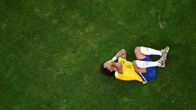 A imagem mostra um jogador da seleção brasileira de futebol caído no gramado, encolhido e com as mãos cobrindo o rosto, sugerindo dor ou frustração após um lance. Registrada de um ângulo aéreo, a fotografia destaca o contraste entre a figura do atleta e a ampla área verde do campo, reforçando a sensação de isolamento no momento.