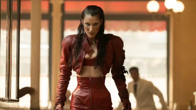 Фотографія Белли Хадід у червоному вбранні з суровим виразом обличчя. Фон розмитий, але можна побачити чоловіка в білій сорочці.