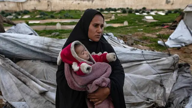 Una madre con un bebé en brazos junto a una tienda de campaña destruida.