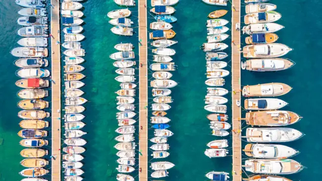 Turkuaz sularda sıralanmış yatların bulunduğu kalabalık bir tekne marinasının havadan görünümü.