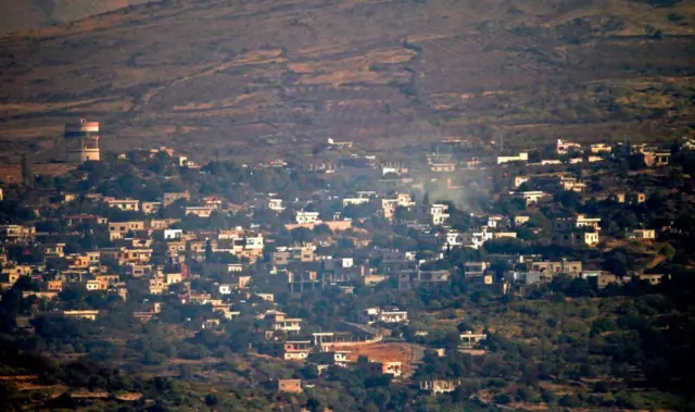 صورة تظهر تصاعد أعمدة الدخان من قرية حضر الدرزية من أعلى تلال خضراء تكسوها مبان أسمنتية صغيرة.