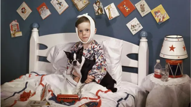 Seorang anak laki-laki sakit dengan perban melilit kepalanya duduk di tempat tidur, dengan anjing bulldog di pangkuannya