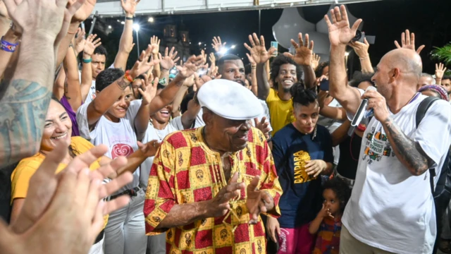 Homem idoso negro reverenciado por público ao fundo