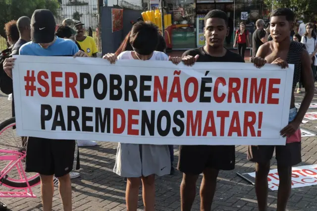 Fotografia mostra adolescentes segurando cartaz que diz "ser pobre não é crime" e "paremslot yg dapat freebetnos matar" 