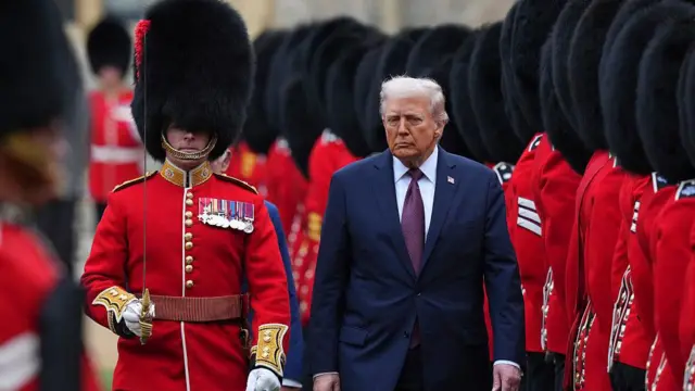 Donald Trump on black suit and burgundy tie, royal guard stand beside am as e carry sword wit full hair. Di royal guards wear red military uniform
