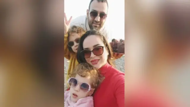 A family photo of Nazanin, her two daughters Deniz and Delsa, and husband Amin in a selfie on a beach with pebbles.