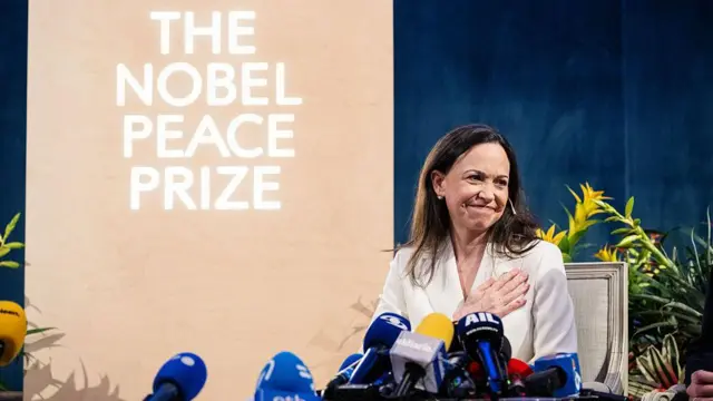 María Corina Machado por trás de microfones, com um cartaz do Prêmio Nobel da Paz ao fundo