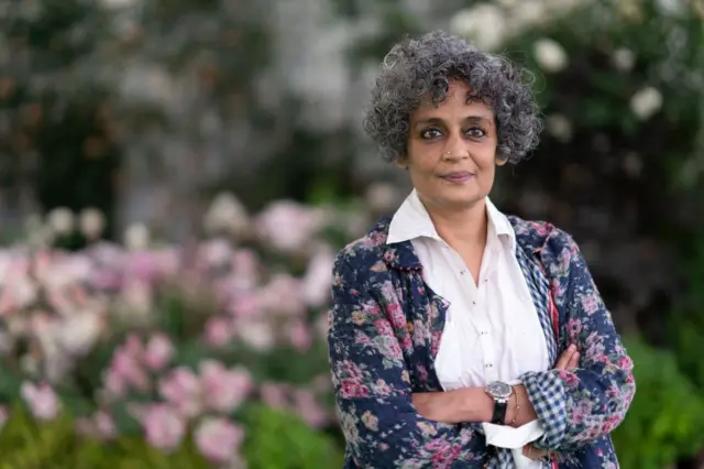Arundhati Roy ellerini birleştirmiş, arkada yeşillik ve çiçekler var. 