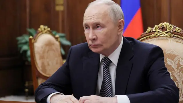 Владимир Путин у тегет оделу за столом гледа у страну, а иза њега се назира руска застава