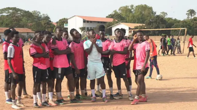 Yankuba Minteh, vêtu d'un maillot d'entraînement bleu clair de Brighton, pose pour une photo aux côtés des membres masculins d'une équipe de la Bakoteh United Football Academy. Ils se tiennent debout sur un terrain sablonneux, vêtus de maillots de football roses et de shorts noirs, tandis que d'autres joueurs transportent un but à l'arrière-plan et que l'on aperçoit au loin des bâtiments bas et des arbres.