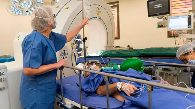 A health worker leans over a patient lying on a hospital trolley with a frame around his head, preparing for brain surgery.