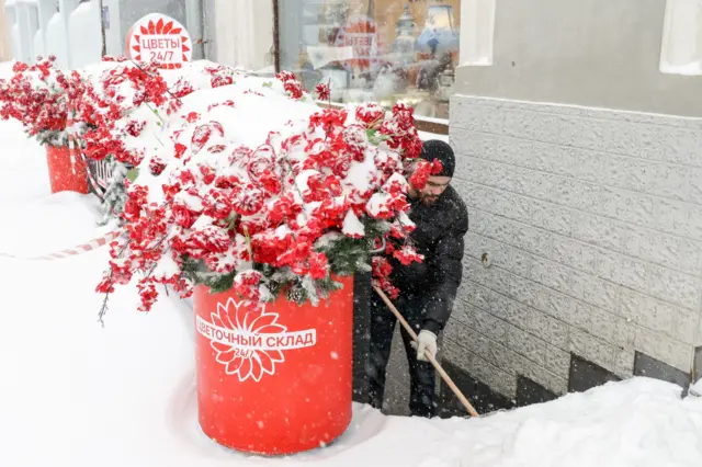 Сотрудник цветочного магазина чистит лестницу, рядом красиво засыпанный снегом вазон с хвойными ветками и ветками красных ягод, похожих на рябину