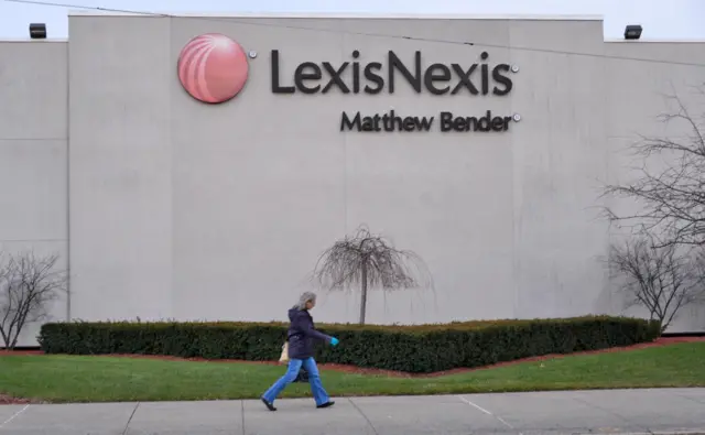 На заднем фоне серая стена здания. На стене - изображение красного шара и надпись LexisNexis Matthew Bender. Перед стеной - деревья без листьев, кусты и газон. На переднем плане по дороге вдоль стены идет человек.