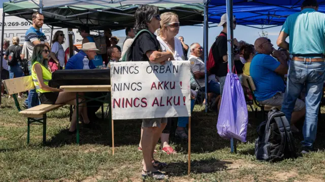 Egy résztvevő „Nincs Orbán, nincs akku, nincs alku!” feliratú táblát tart a Tisza párt szabadtéri rendezvényen sátor alatt ülő emberek mellett.