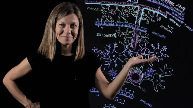 A professora Noelia Valle com camiseta preta e cabelos loiros, em frente a uma lousa com desenhos coloridos sobre biologia