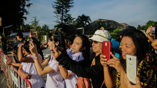 Mulheres tiram fotos com celulares ao ladovbet giros grátiscerca que protege acampamento perto da caverna onde meninos ficaram presos