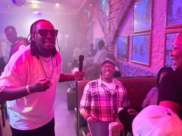 Jesus Marie Joseph Kavyinabuhiye afise micro mu kuboko kw'ibumbafu hafi hari umugabo yambaye ishati ya karokaro biboneka ko yaryohewe cane. 