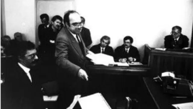 Uğur Mumcu bir mahkeme salonunda sanık veya tanık kürsüsünde ayakta konuşurken görülüyor. Arkada avukatlar, mahkeme heyeti üyeleri ve izleyiciler bulunuyor. 