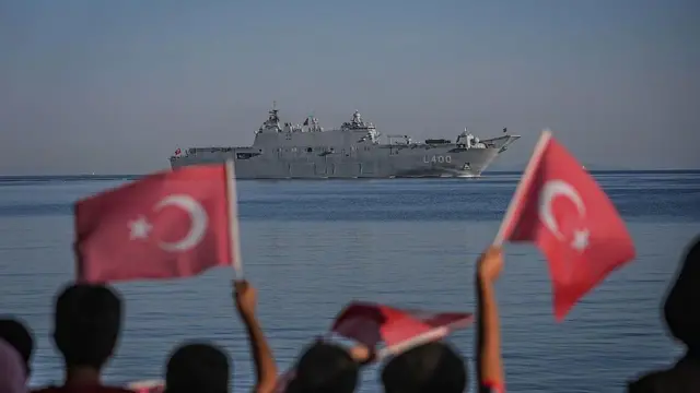 Çocukların flu bayran sallayan görüntüleri önde, hepsinin arkası dönük denizden geçen gemiye bakıyorlar.