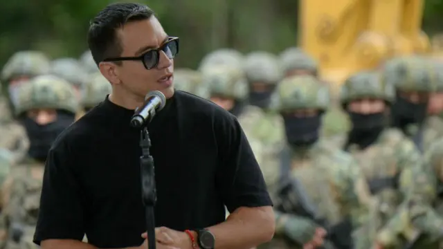 Ekvador Devlet Başkanı Daniel Noboa, arkasında silahlı askerler varken bir mikrofonun önünde konuşuyor. Kısa siyah saçlı olan Noboa, siyah güneş gözlüğü ve siyah bir tişörtü var. 