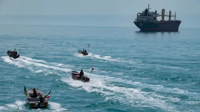 Cuatro lanchas rápidas con banderas iraníes navegando por el Golfo Pérsico, cerca de un buque de carga general.