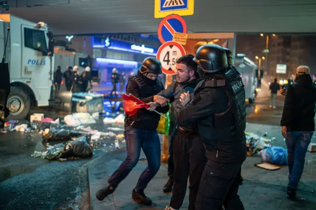 İki polis elinde bayrak tutan bir kişiyi kollarından tutarken görülüyor. 
