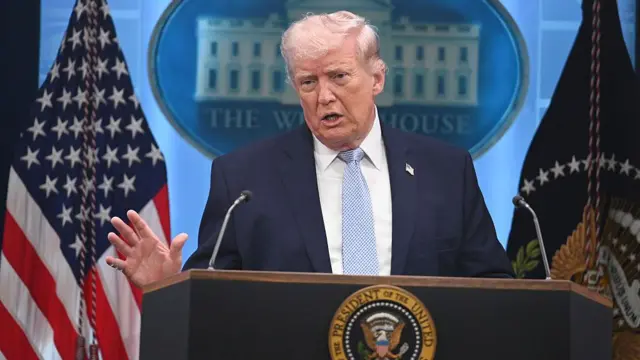 O presidente americano, Donald Trump, com terno azul escuro e gravata azul clara, de pé, atrás de um pódio.
