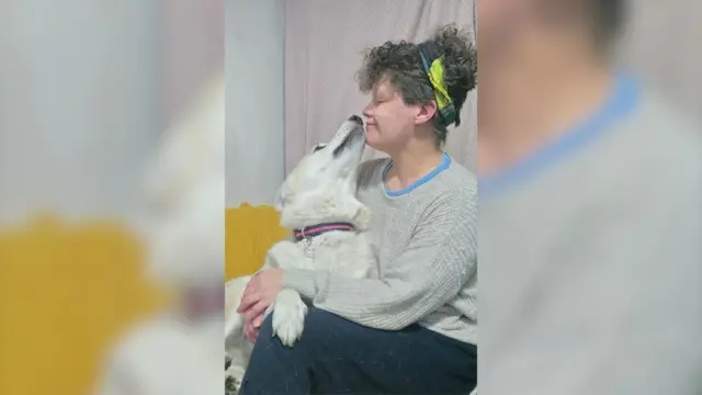 Una mujer abraza a su perro. Están frente a frente y el hocico del perro roza el suyo. El perro es blanco con un collar rojo y negro, y la mujer tiene el pelo castaño y rizado.