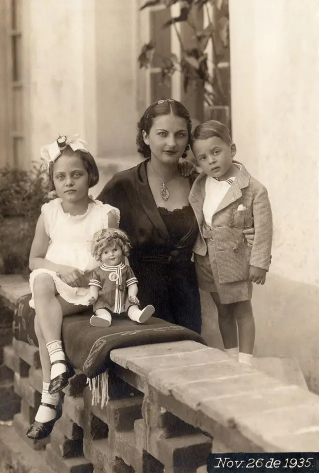 Cristina, en el centro, lleva un vestido y una blusa negros y un collar. A su lado está Antonio, quien lleva una camisa blanca y una chaqueta y un pantalón corto color gris. Al otro lado, sentada, está Isolda, que viste un traje blanco sin mangas, medias blancas y zapatos negros y un lazo en la cabeza. Una muñeca está sentada delante de ellas. Están en un jardín.