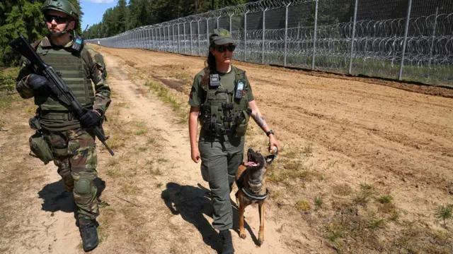Dwóch żołnierzy, kobieta i mężczyzna, idzie z psem wzdłuż suchej, błotnistej ścieżki obok dużego ogrodzenia z drutu ostrzowego. W tle widać las. Żołnierz niesie karabin, a żołnierka trzyma zabawkę dla psa.