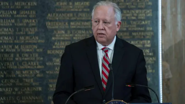 Thomas Shannon com terno escuro, camisa branca e gravata listrada fala em púlpito. Ele é branco e tem cabelos brancos; ao fundo, há um monumento com vários nomes de pessoas