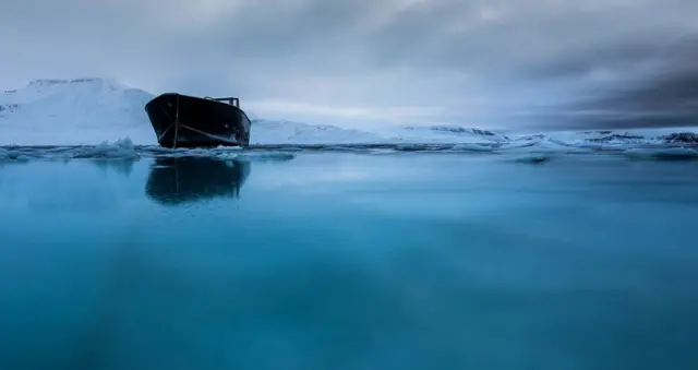 Mali čamac na plavoj vodi sa planinama u pozadini. Fotokrafisao Kler Kajns