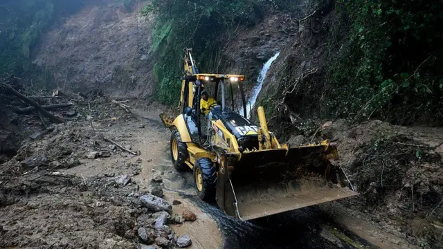 Un hombre con chaleco amarillo conduce un tractor amarillo para despejar el lodo y las rocas de una carretera en una montaña