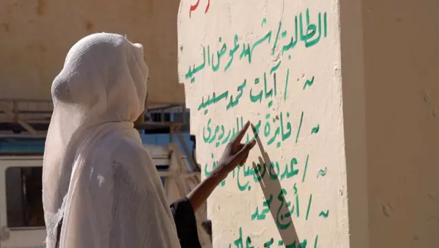 مكارم أمام جدار كُتب عليه أسماء الطالبات اللاتي قتلن في الهجوم.