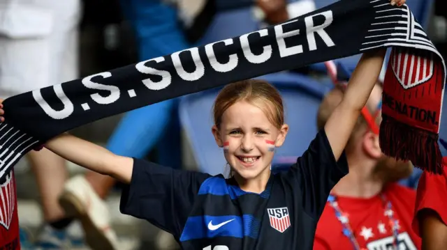 Una niña con una bufanda negra que dice U.S. Soccer en letras blancas. Lleva una camisa de fútbol de color azul, con el número 16 y el escudo que dice USA.
