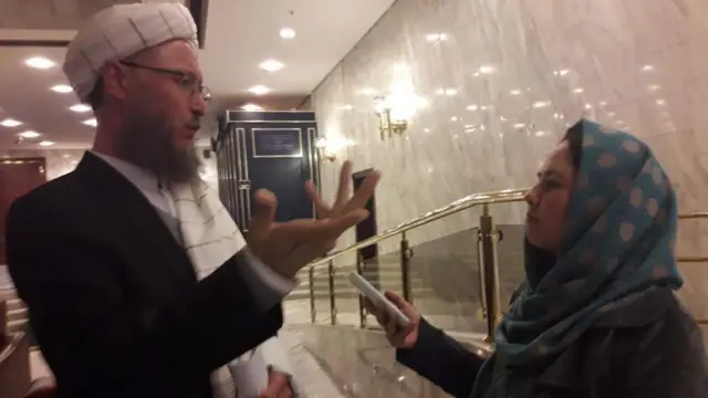 A slightly blurry image showing Zahra holding out a recording device or smartphone towards a man who is smartly dressed in clerical clothing, with a black jacket and white turban. He is gesticulating with his hand. She is wearing a blue and white polka dot headscarf.