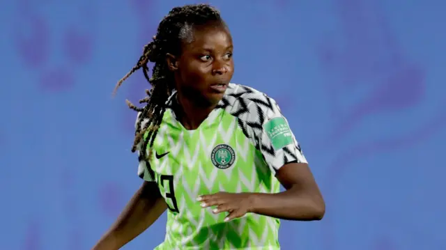 Ngozi Okobi-Okeoghene, visible jusqu'à la taille, vêtue d'un maillot vert et blanc du Nigeria avec des bordures noires et blanches sur le haut des bras, lève le bras gauche à hauteur de poitrine et regarde vers la gauche pendant un match de football.