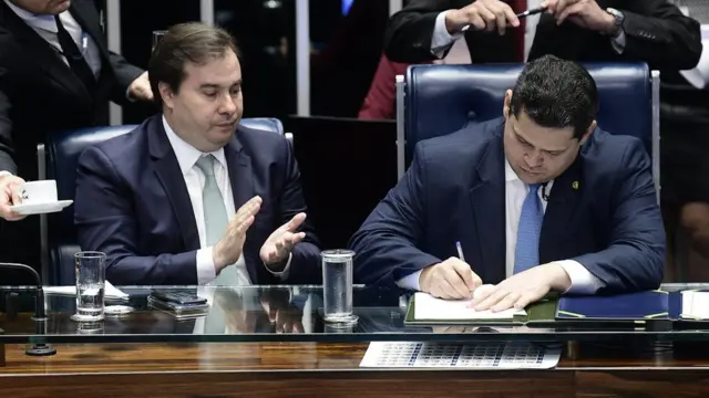 No Congresso, Rodrigo Maia e Davi Alcolumbre aparecem sentados à mesa - o primeiro batendo palmas e o segundo, assinando papel