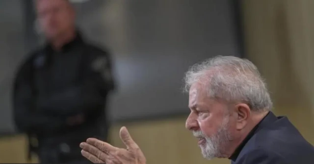Imagem mostra Lula, de perfil, usando um terno cinza e falando a uma repórter com um policial federal desfocado no fundo da imagem