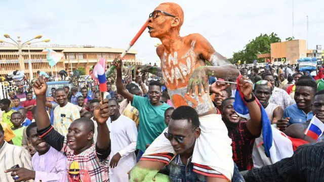 Abantu barimo n'abaseka bafite amabendera, n'umugabo uhetse undi ku rutugu