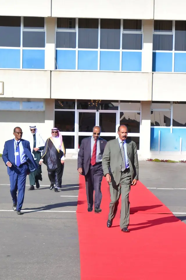 ወግዓዊ ልብሲ ዝተኸደ ፕረዚደንት ኢሳይያስ ኣፈወርቂ  ኣብ ቀይሕ ምንጻፍ ናብ ነፋሪት እናሰጎመ። ብድሕሪኡ፡ መነጸር ዓይኒን ቀይሕ ካራቫታ ዝገበረ ሚኒስተር ዑስማን ሳልሕ እግሪ እግሩ እናሰዓበ፡ ካብ ምንጻፍ ወጻኢ፡ ክልተ ናይ ስዑዲ ባህላዊ ክዳን ዝገበረን፡ ሓደ ሓለዋ ወይ ብዓልመዚ ፕሮቶኮል ካብ ምንጻፍ ወጻኢ እናሰዓብዎም።