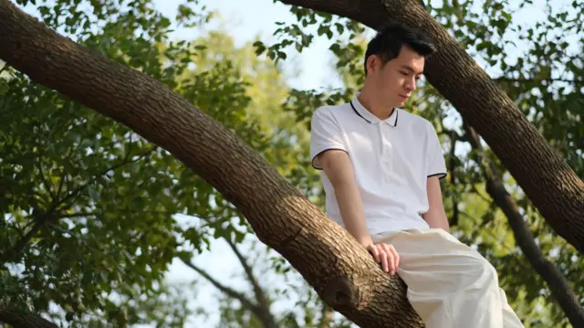 A man wearing white clothes sits on a tree branch, looking pensively down towards the ground.