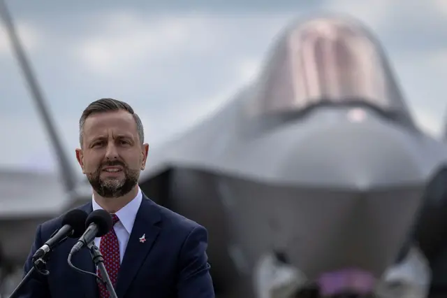 Косіняк-Камиш в діловому костюмі виступає біля мікрофонів на тлі сучасного військового літака. Знімок зроблено просто неба під час офіційного заходу, пов’язаного з авіацією або оборонною сферою.