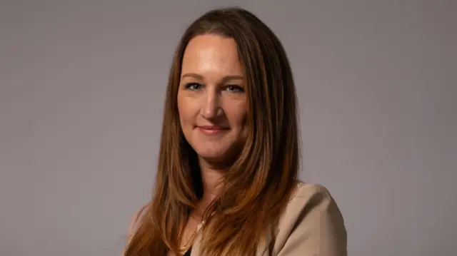 Kejt, elegantno obučena žena u bež odelu, sa dugom smeđom kosom, osmehuje se kameri. 