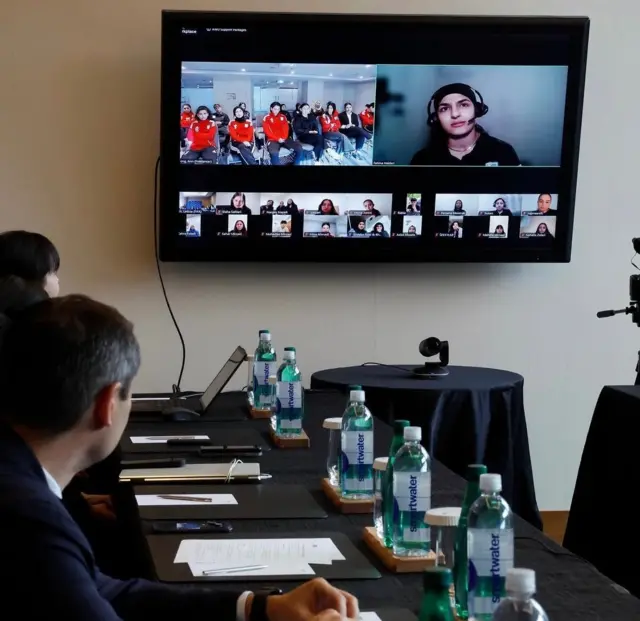 په کوټه کې ځينې چارواکي‌ ناست دي چې پر میز یې دوسیې او د اوبو بوتلونه ایښي، دوی پر دیوال لګېدلي سکرین ته ګوري چې د افغان ښځينه لوبډلې غړې پکې انلاین برېښي.