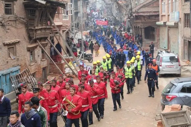 भूकम्प सुरक्षा दिवसको र्‍याली, छेउमा भूकम्पपछि जीर्ण बनेको पुरानो भवनमा झ्याललाई टेको लगाइएको देखिन्छ 