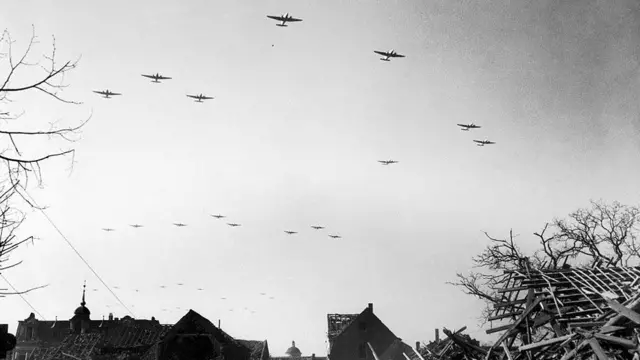 Allied planes fly over a shattered German town on their way to drop paratroopers over the Rhine in March 1945. The black and white images shows planes in the sky. Below are roofs with their struts showing, and on the right is a heap of roof struts exposed to the sky. On both sides are leafless tree branches.