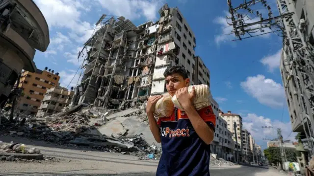 Um jovem palestino carregando pão na Cidadeesporte bet youGaza, caminhando entre prédios destruídos