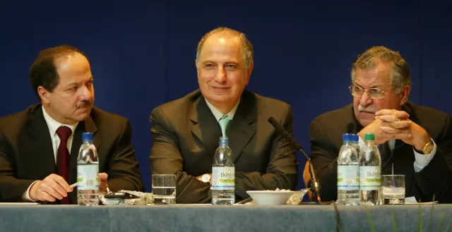 Üç siyasetçi takım elbise giyiyor, önlerinde su ve bardaklar var. Çelebi önünde mikrofon var.